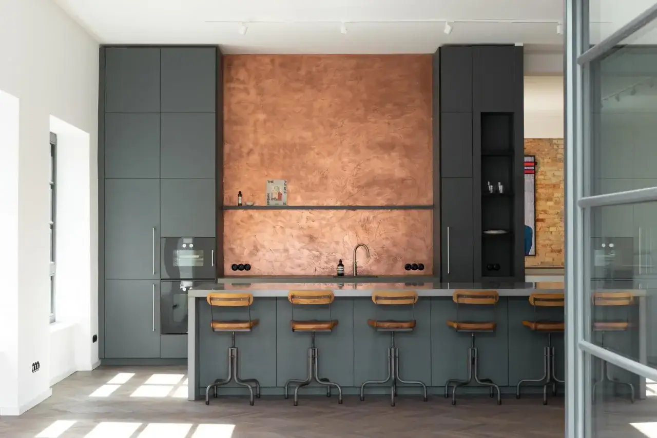 Cuisine moderne avec mur texturé en laiton cuivre, meubles gris anthracite et tabourets industriels.