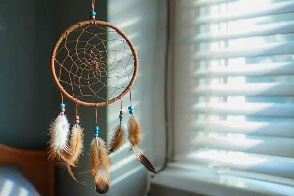 Attrape-rêves artisanal avec plumes et perles bleues, suspendu devant des stores.