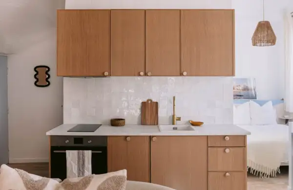 Petite cuisine de studio avec placards en bois, carrelage blanc et lit en arrière-plan.
