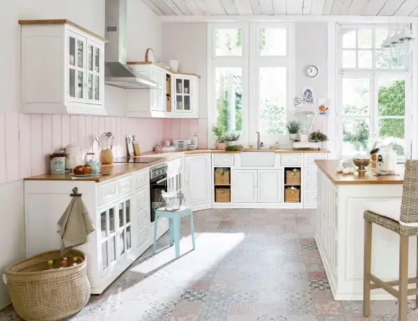 Une cuisine blanche style campagne chic, lumineuse avec ses grandes fenêtres, son plan de travail en bois et son sol aux motifs colorés.