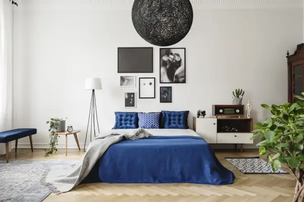 Chambre bleu nuit avec lit, coussins capitonnés, plaid gris, lampadaire, table de chevet et décoration murale.