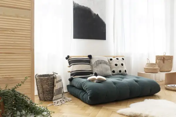 Futon vert foncé avec coussins décoratifs, panier en osier et tableau abstrait.