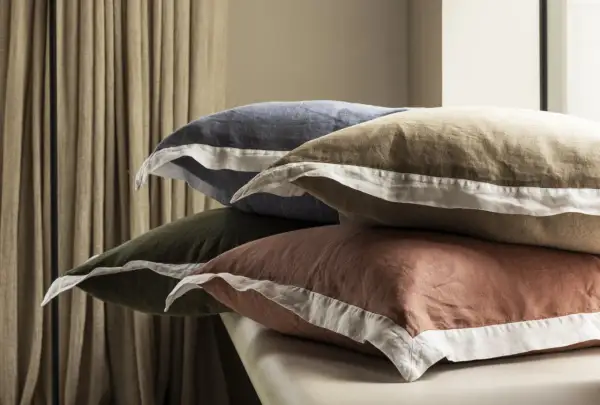 Pile de taies d'oreiller de différentes couleurs (bleu, kaki, marron) avec passepoil blanc.